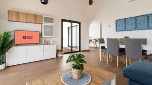 Ein modernes Wohnzimmer mit Fernseher, einer Essecke mit Stühlen, einem großen Fenster und einer Pflanze auf einem Holztisch.