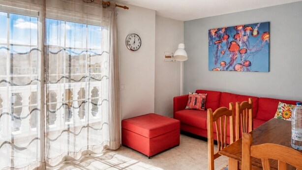 A room with a red sofa, a wooden dining table, a clock on the wall, and a painting above the sofa. - GuestReady - Arenas Charming Pool (Caleta de Fuste)