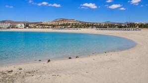 A beach with clear turquoise water, a sandy shore, and a backdrop of buildings and a hill. - GuestReady - Arenas Charming Pool (Caleta de Fuste)