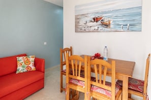 A room with a red sofa, wooden dining table with chairs, and a painting of boats on the wall. - GuestReady - Arenas Charming Pool (Caleta de Fuste)