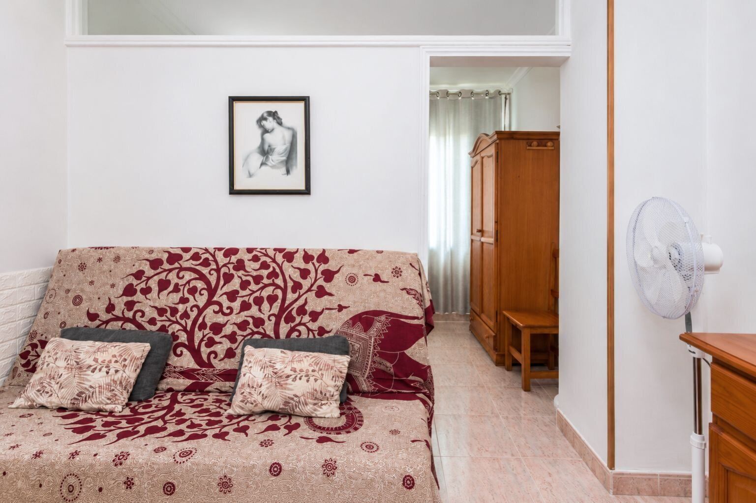 Une pièce avec un canapé à motifs, un meuble en bois, un ventilateur et un tableau encadré au mur.