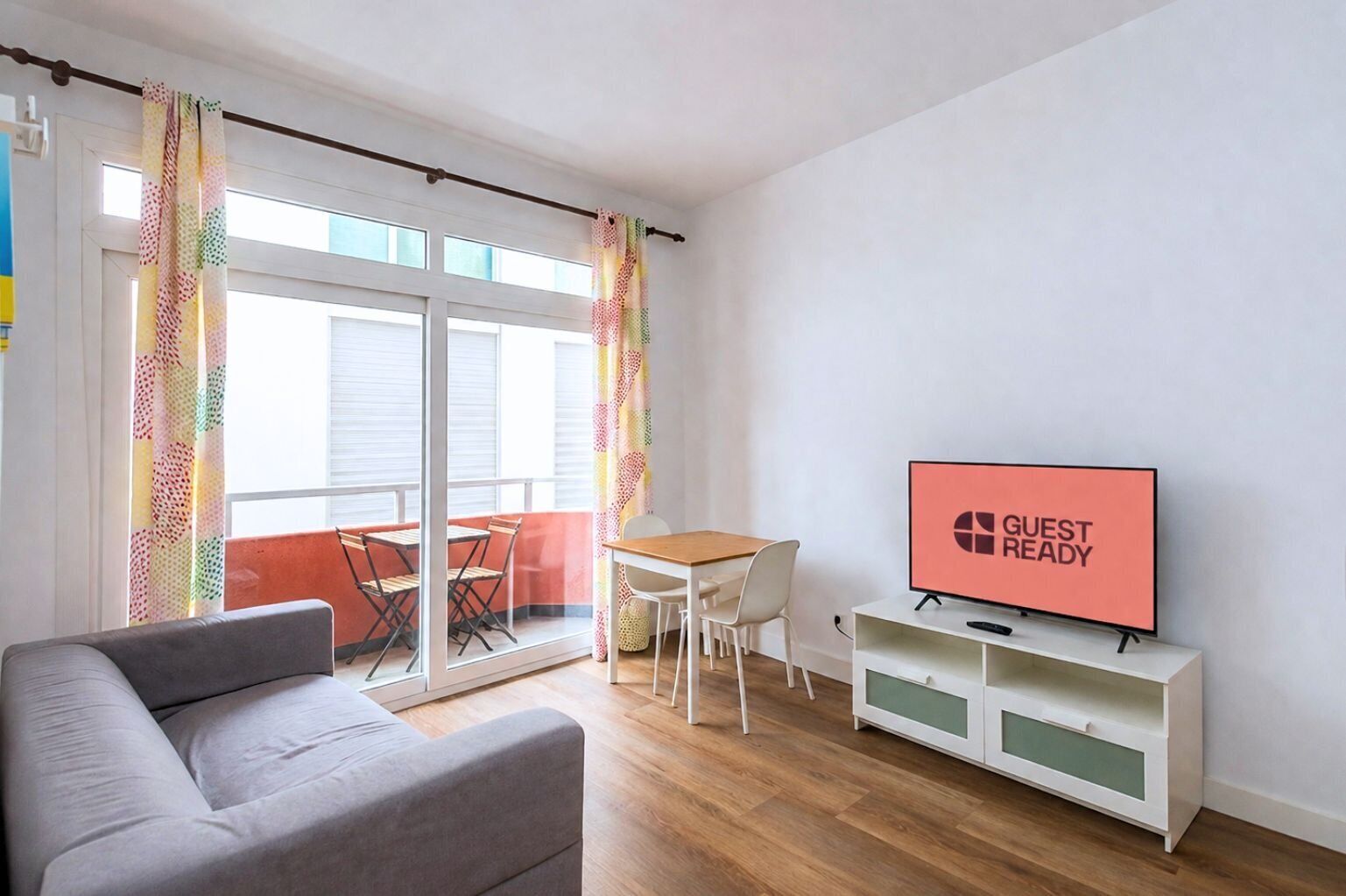 Una sala de estar moderna con un sofá, un televisor mostrando 'GUEST READY', un balcón con mesa y sillas, y un ventanal corredero.