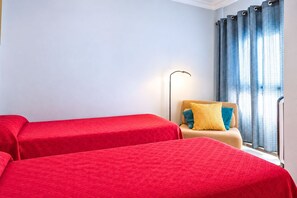 Una habitación de hotel con dos camas, un sofá con cojines, una lámpara de pie y una ventana con cortinas.