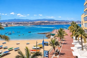 Une vue côtière avec des bateaux sur la plage, un ciel dégagé et des montagnes à l’horizon.