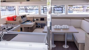Interior de barco com cozinha, painel de controle, área de estar com mesa e cadeiras, e janelas com vista para a marina.