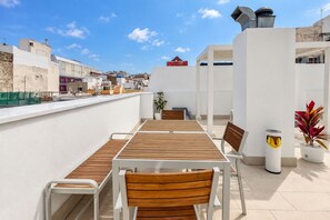 Terraço ao ar livre com mesa e cadeiras de madeira, uma planta em vaso e uma lixeira.