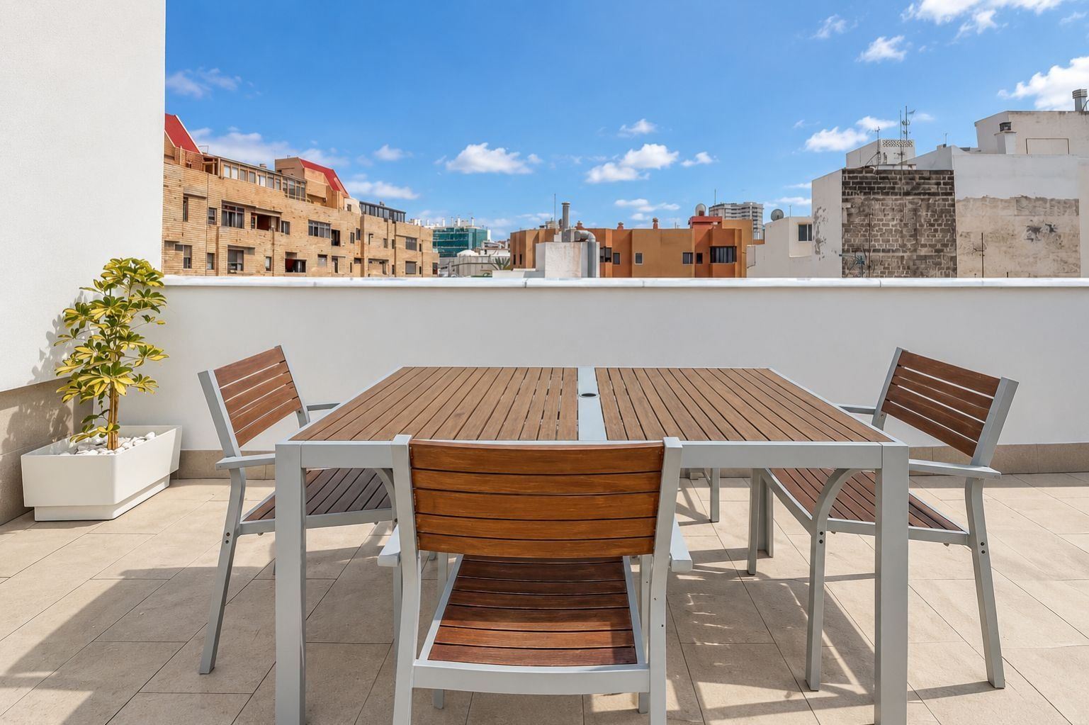 A rooftop terrace with a wooden table and chairs, a potted plant, and a city view.