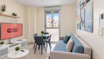 A modern living room with a glass dining table, a sofa, a TV displaying 'GUEST READY', and a city view through the window.