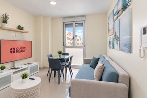 Una sala de estar moderna con una mesa de comedor de vidrio, un sofá, un televisor mostrando 'GUEST READY' y vista a la ciudad a través de la ventana.