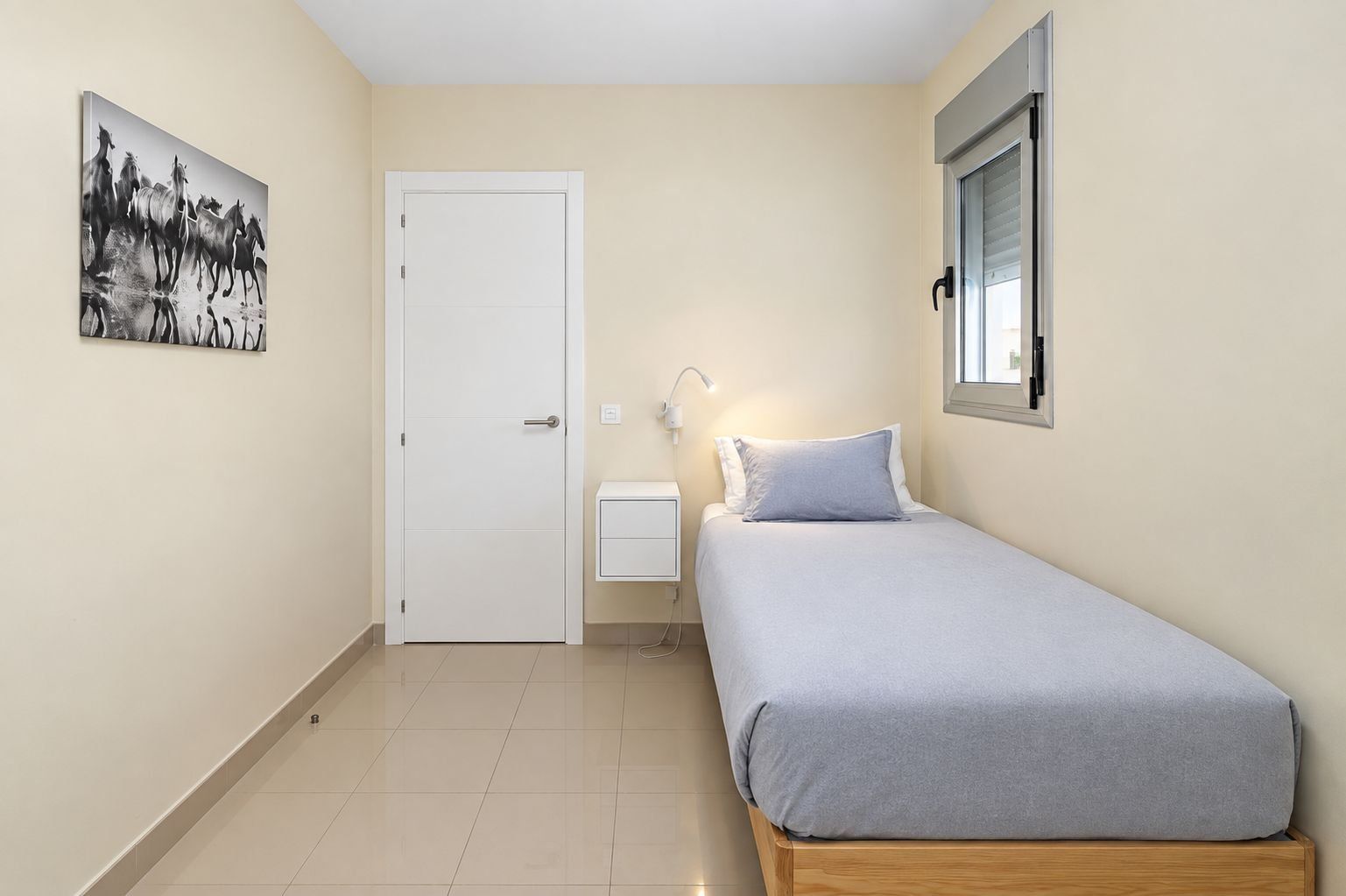 Une chambre à coucher comprenant un lit, une table de chevet, une commode, une lampe fixée au mur, une porte et une fenêtre.
