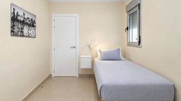 A bedroom with a bed, a bedside table, a nightstand, a wall-mounted lamp, a door, and a window.