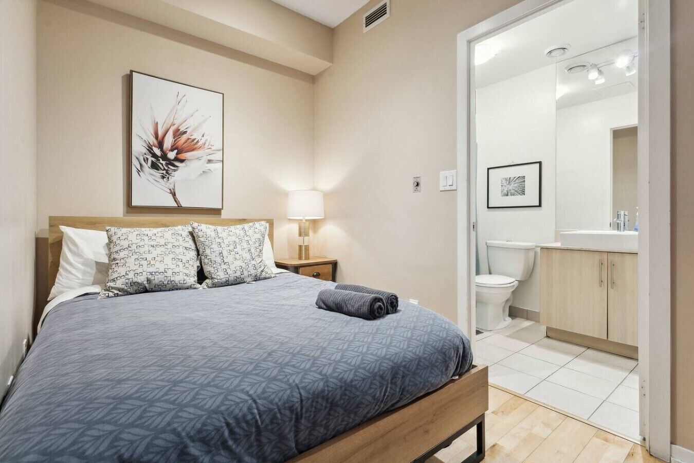 A bedroom with a bed, pillows, a bedside lamp, a framed picture on the wall, and a bathroom visible through an open door.