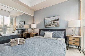 Hotellihuoneessa on suuri sänky, pienempi sänky, yöpöytä, jossa on lamppu, peili ja taulu seinällä.