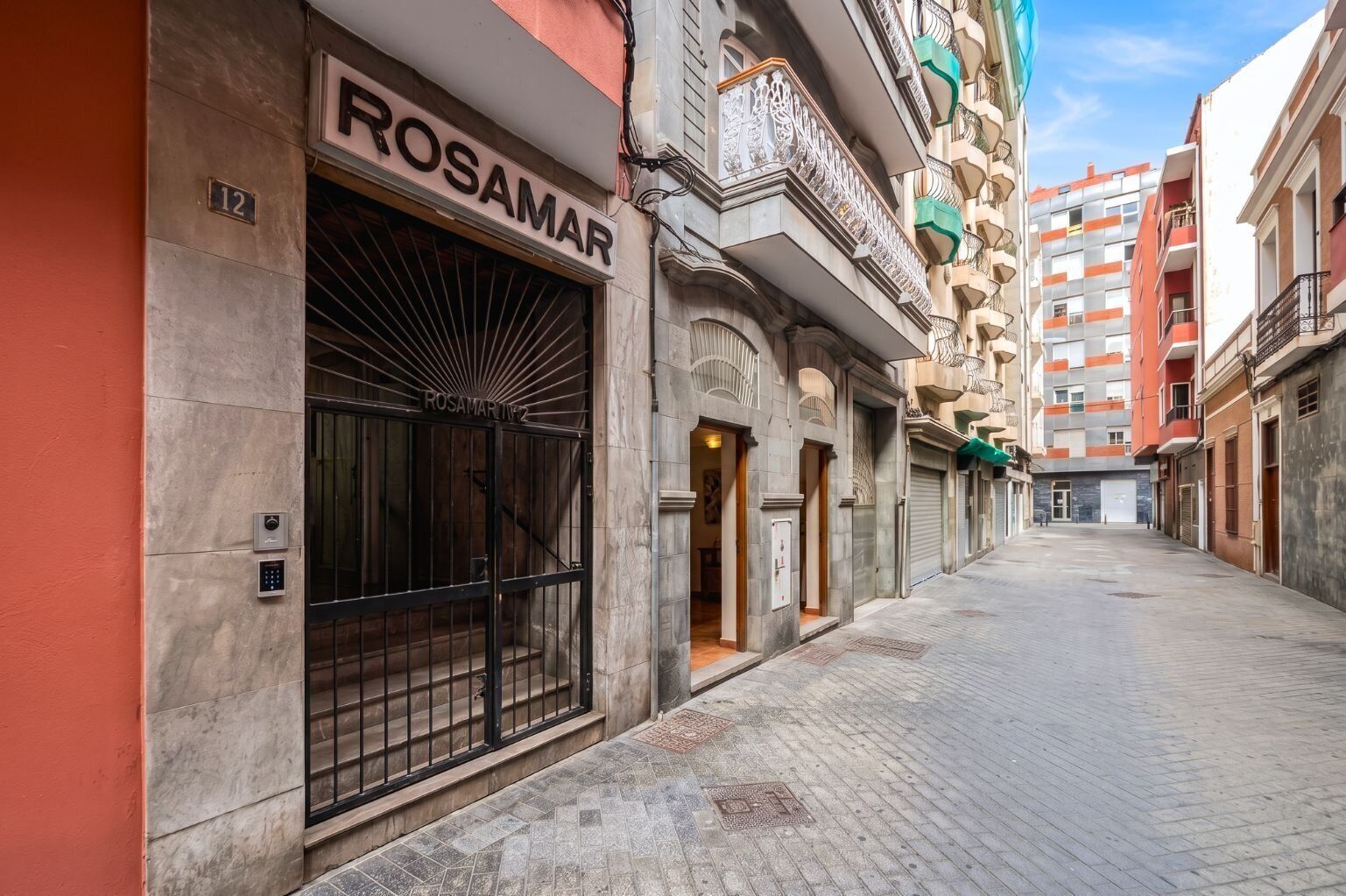 Una calle estrecha con un edificio identificado como 'ROSAMAR' a la izquierda, que presenta una gran puerta metálica y una entrada pequeña.