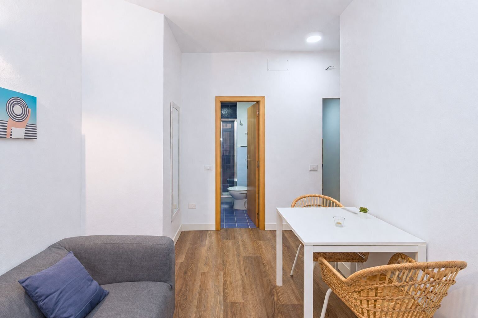 Una habitación pequeña y moderna con un sofá, una mesa de comedor y una silla. Se ve una puerta que da a un baño con inodoro y ducha.
