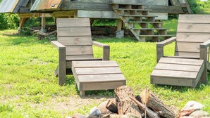 A rustic wooden cabin with a triangular roof, a small attached shed, and a fire pit with logs.