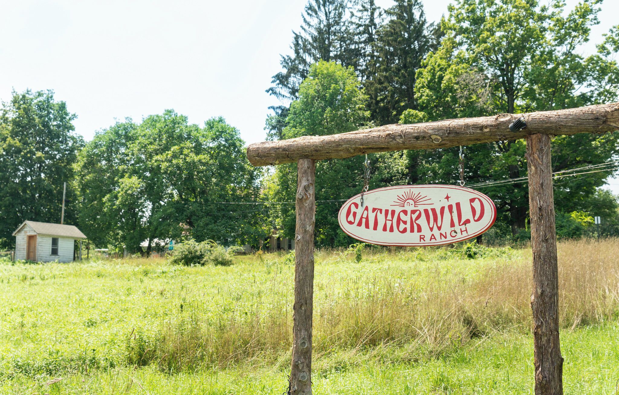 Un letrero de madera rústico con la inscripción 'GATHERWILD RANCH' en un entorno rural, con una casa pequeña y árboles densos al fondo.