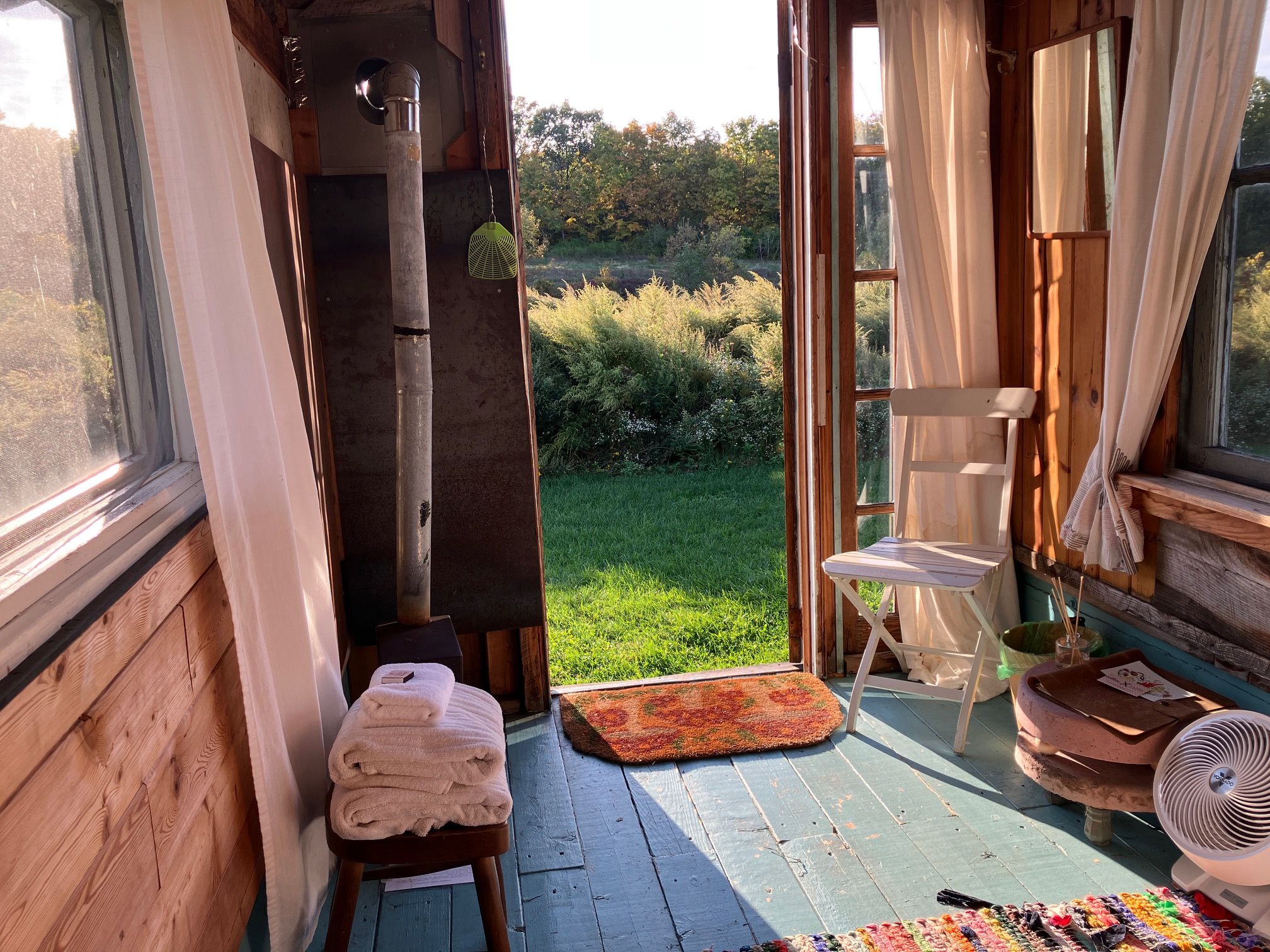 Une pièce en bois donnant sur un espace verdoyant, avec un petit tabouret, un ventilateur et une table recouverte d’un tapis.