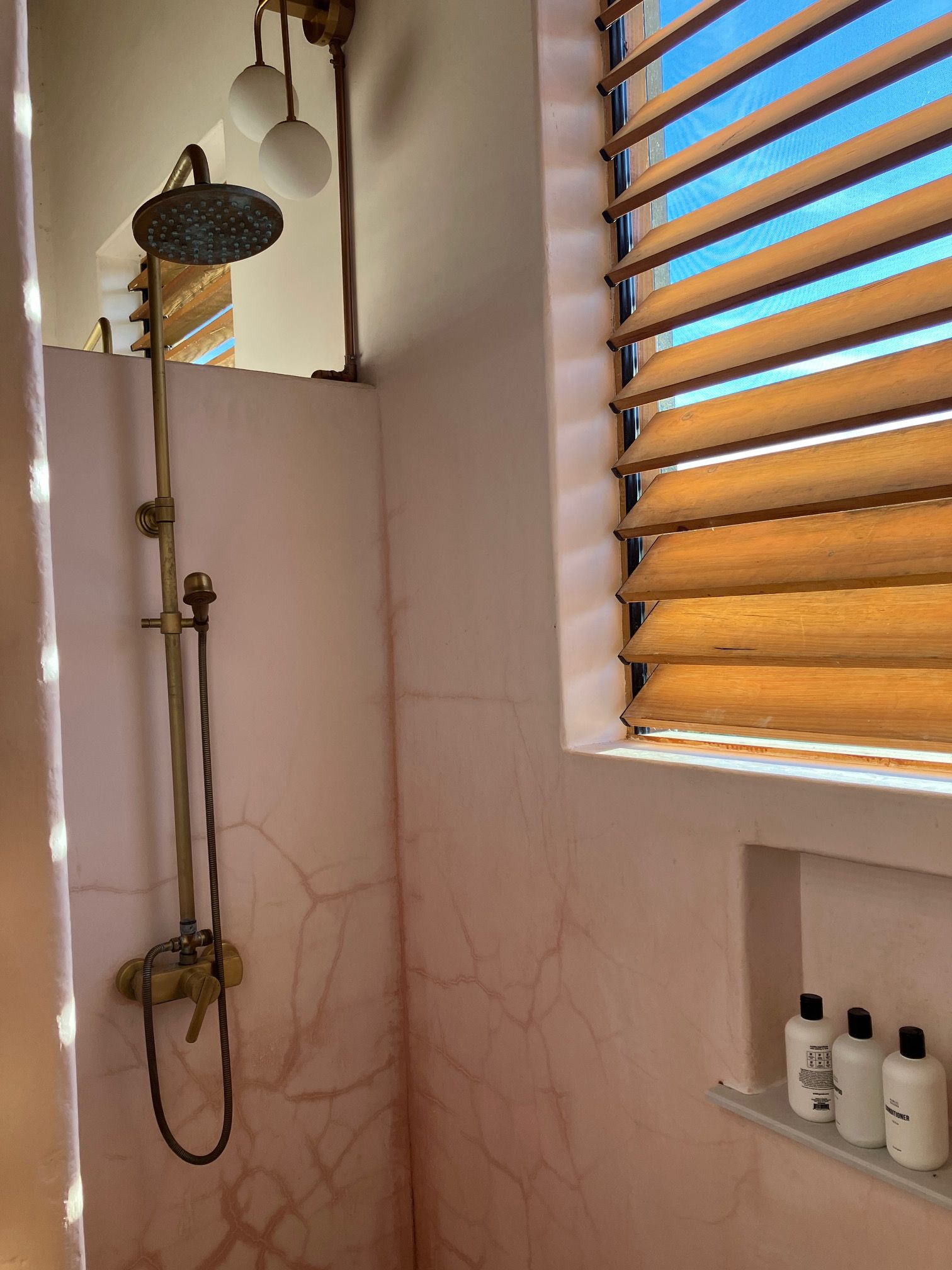 Un baño con ducha, una ventana con persianas de madera y un estante con artículos de aseo.