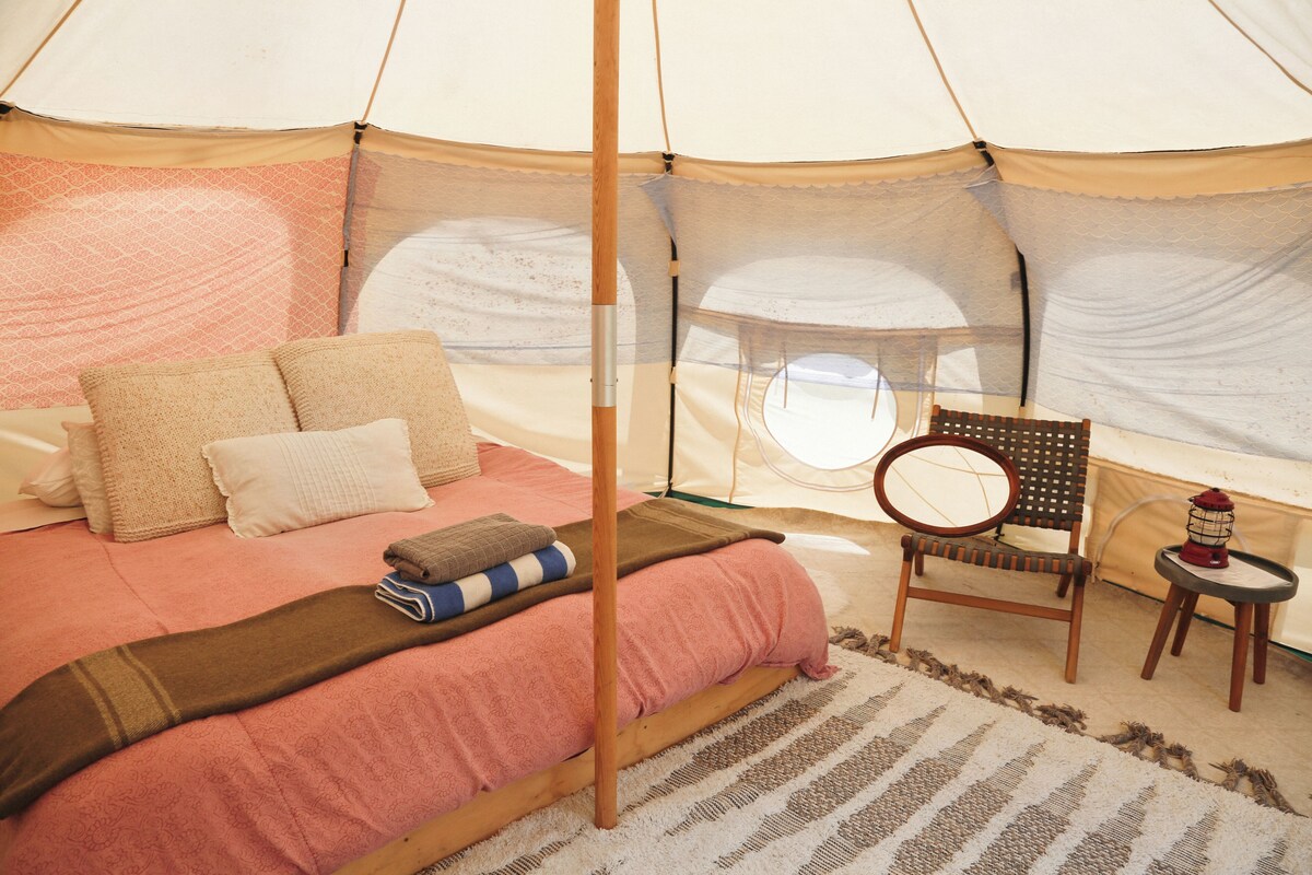 A tented room with a bed, pillows, a chair, a small table, and a lantern.