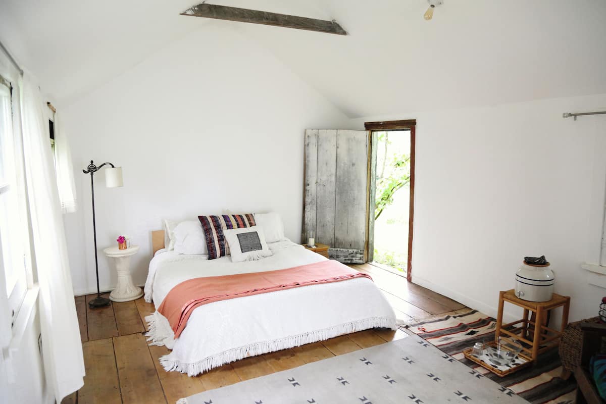 A bedroom with a bed, bedside table, lamp, and a view of the outdoors.