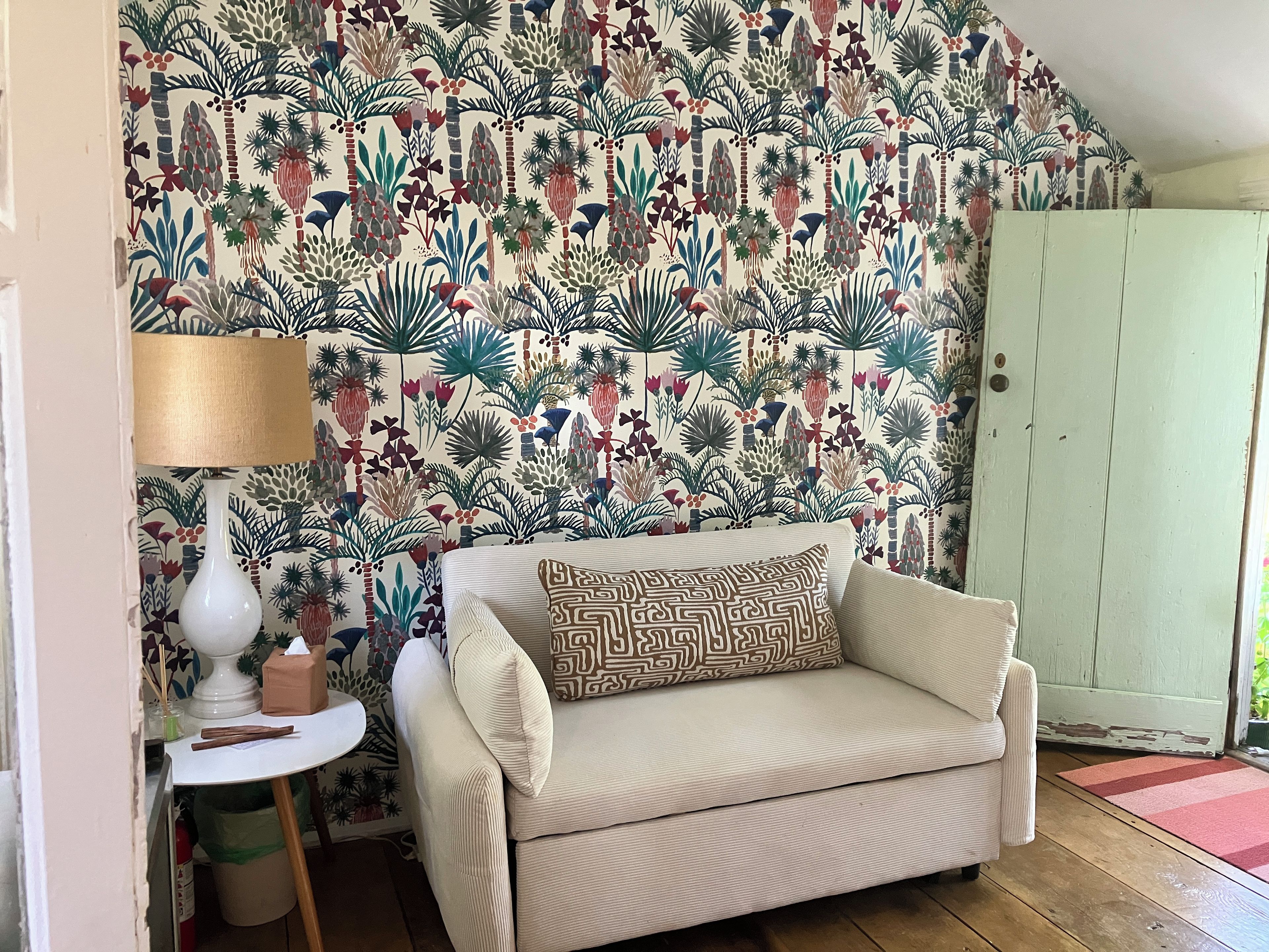 A room with a patterned wallpaper, a beige sofa with a patterned pillow, a small white table with a lamp, and a wooden floor.