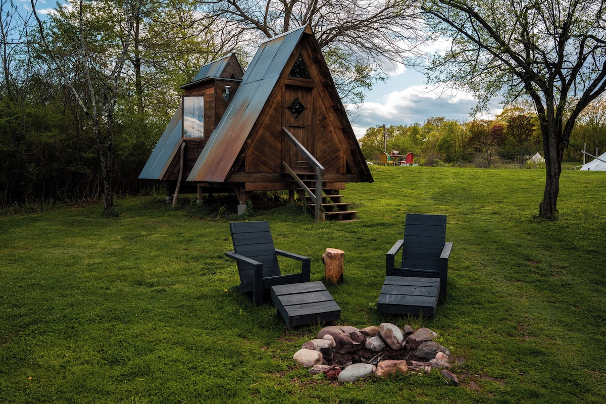A wooden cabin with a triangular roof, a fire pit, and two Adirondack chairs.