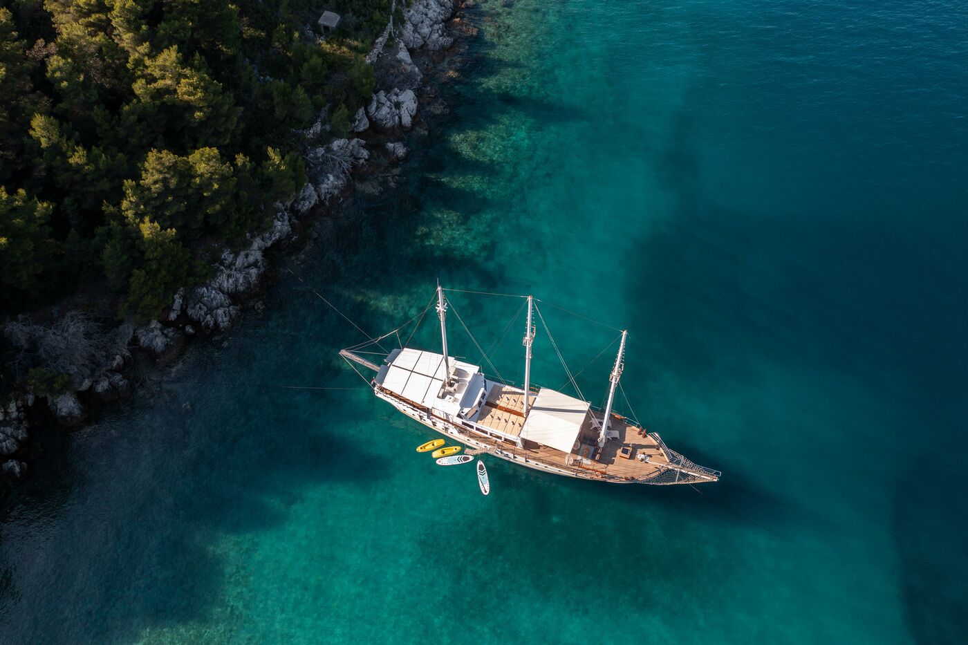 Une vue aérienne d’un voilier ancré dans des eaux turquoise et limpides, entouré d’une côte rocheuse et d’une végétation luxuriante.