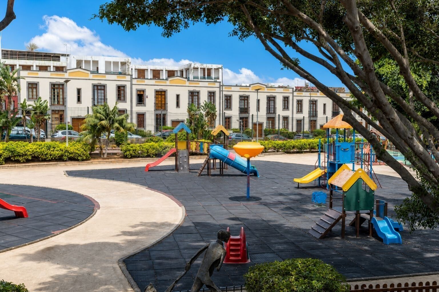 A playground with various play structures, surrounded by a paved area and a building in the background.
