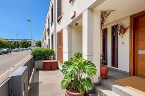 Ein Gebäude mit überdachter Eingangspartie, ein Blumentopf im Vordergrund und eine Straße mit parkenden Autos.