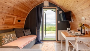Una cabaña acogedora con interior de madera, un sofá con cojines estampados, una mesa de comedor con una botella de vino y vistas a través de un gran ventanal.