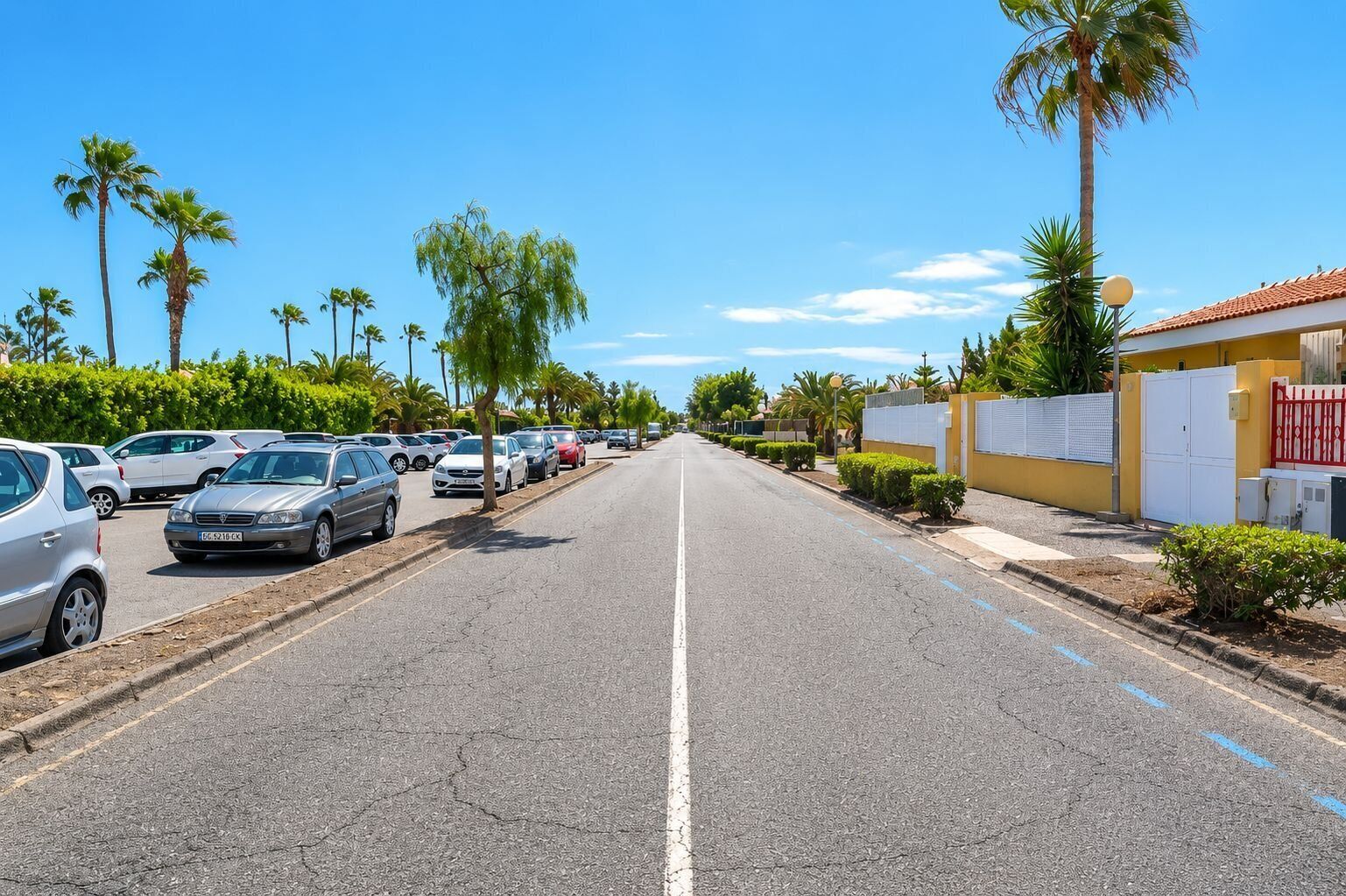 Rua com carros estacionados e palmeiras, que leva a um prédio com fachada amarela e portão vermelho.