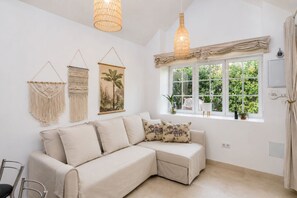 A modern living room with a beige sectional sofa, macrame wall hangings, and a large window with a view of greenery. - GuestReady - Los Tunos BBQ Garden (San Bartolomé de Tirajana)