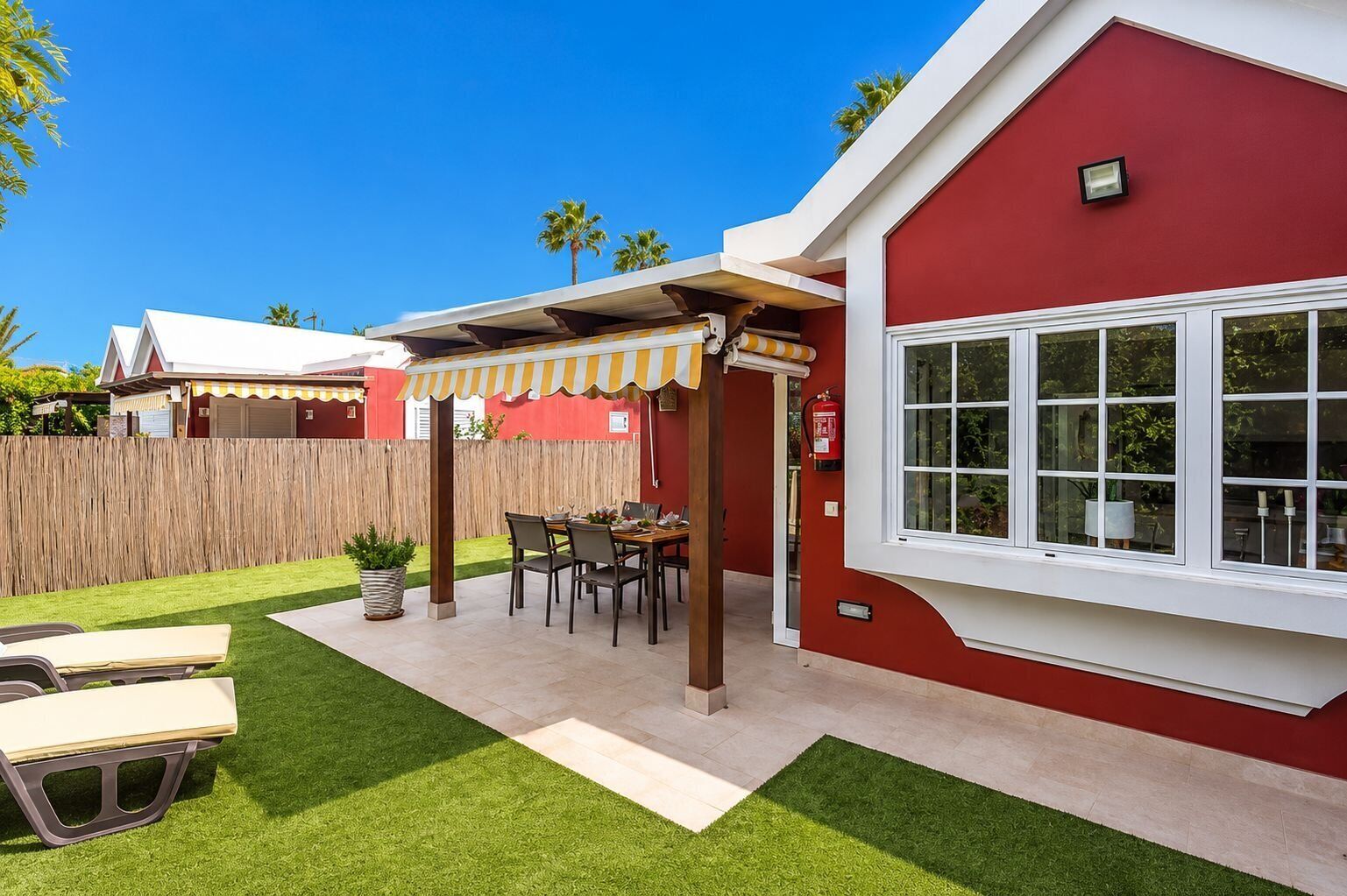 A red building with a white window, a covered patio with a table and chairs, and a wooden fence in the background.