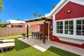A red building with a white window, a covered patio with a table and chairs, and a wooden fence in the background. - GuestReady - Los Tunos BBQ Garden (San Bartolomé de Tirajana)
