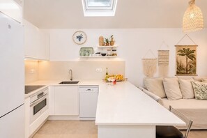 A modern kitchen with white cabinetry, a dishwasher, a built-in oven, a sink, and a dining area with a white table and a sofa. - GuestReady - Los Tunos BBQ Garden (San Bartolomé de Tirajana)