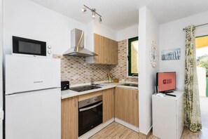 A compact kitchen with a microwave, stove, and sink. There is a small refrigerator and a television on a stand. The kitchen has wooden cabinets and a patterned backsplash. - GuestReady - Cosy Bungalow with Pool and Terrace (Costa Calma)