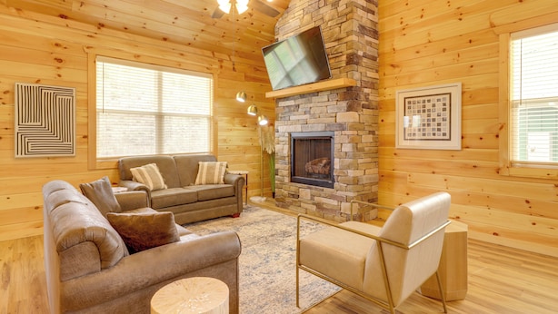 A log cabin interior with a stone fireplace, wooden walls, a sofa, and a television mounted on the fireplace. - Dolly's Cabin: Private Hot Tub, Walk to Dollywood (Pigeon Forge)