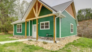 A green house with a porch, stone foundation, and a small table. - Dolly's Cabin: Private Hot Tub, Walk to Dollywood (Pigeon Forge)