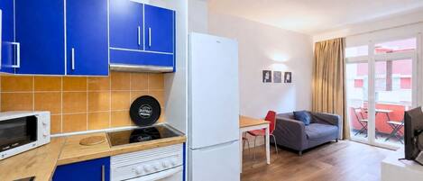 A compact kitchen with blue cabinets, a white refrigerator, a microwave, and a stove. There's a dining area with a table and chairs, and a sofa.