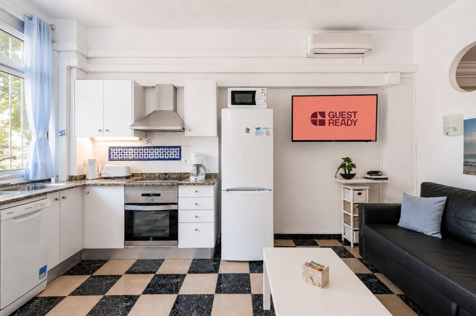 Eine kleine Küche mit weißen Schränken, einer Mikrowelle, einer Kaffeemaschine und einem Kühlschrank. An der Wand hängt ein Schild mit der Aufschrift 'GUEST READY', ein kleiner Tisch mit einer Pflanze und ein schwarzes Ledersofa.