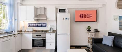 Eine kleine Küche mit weißen Schränken, einer Mikrowelle, einer Kaffeemaschine und einem Kühlschrank. An der Wand hängt ein Schild mit der Aufschrift 'GUEST READY', ein kleiner Tisch mit einer Pflanze und ein schwarzes Ledersofa.