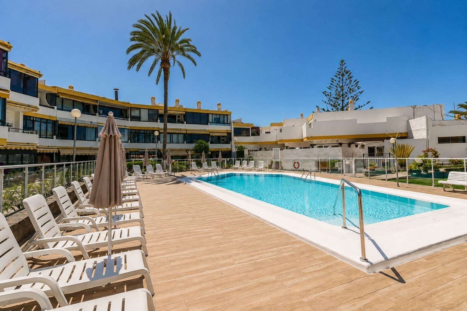 Un espace piscine doté de chaises longues et de parasols, entouré d’immeubles avec balcons.