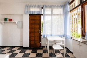 A room with a checkered floor, a wooden door, a white table with chairs, a window with curtains, and a bookshelf. - GuestReady - San Agustin Pool Escape (Playa del Ingles)