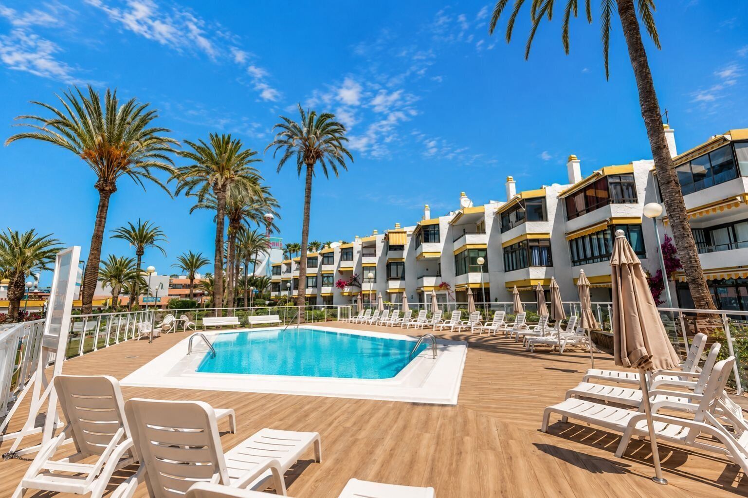 A pool area with lounge chairs and umbrellas, surrounded by palm trees and a multi-story building with balconies.