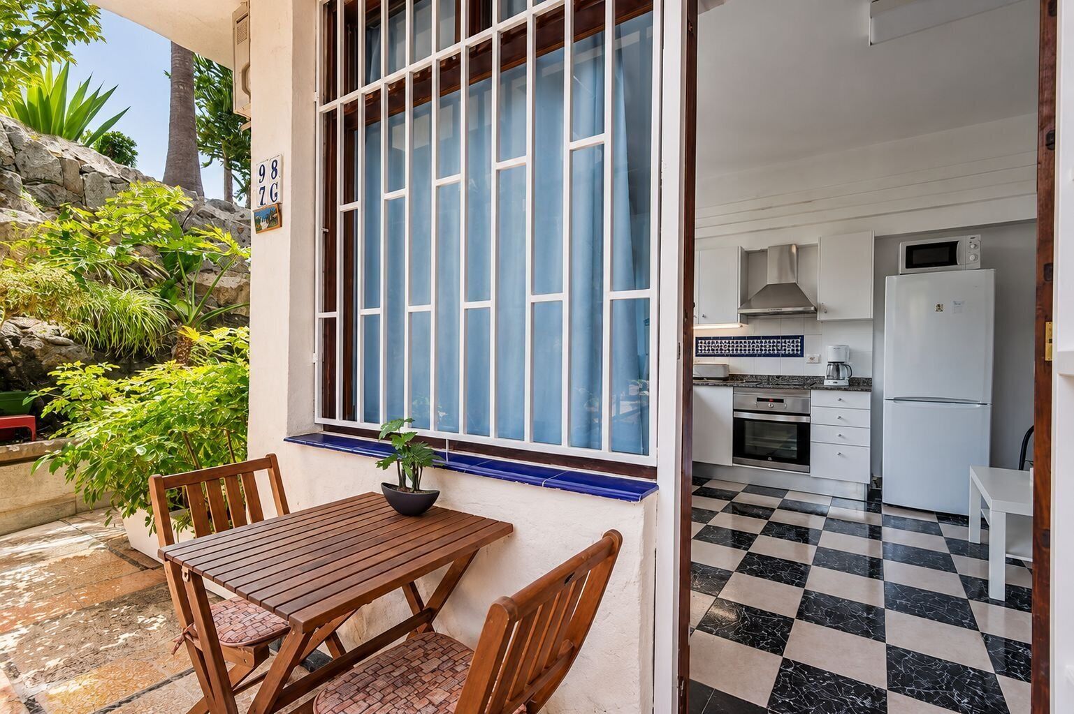 Un patio con una mesa y sillas de madera, una planta en maceta y una ventana con cortinas azules.