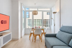 A modern living room with a grey sofa, a dining area with a glass table and white chairs, a TV on the left, and a large window with a view of buildings. - GuestReady - Sunset Canteras Sea Views (Las Palmas de Gran Canaria)