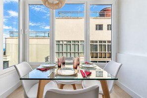 Un comedor con una mesa de vidrio, sillas blancas y vistas a edificios a través de amplios ventanales.