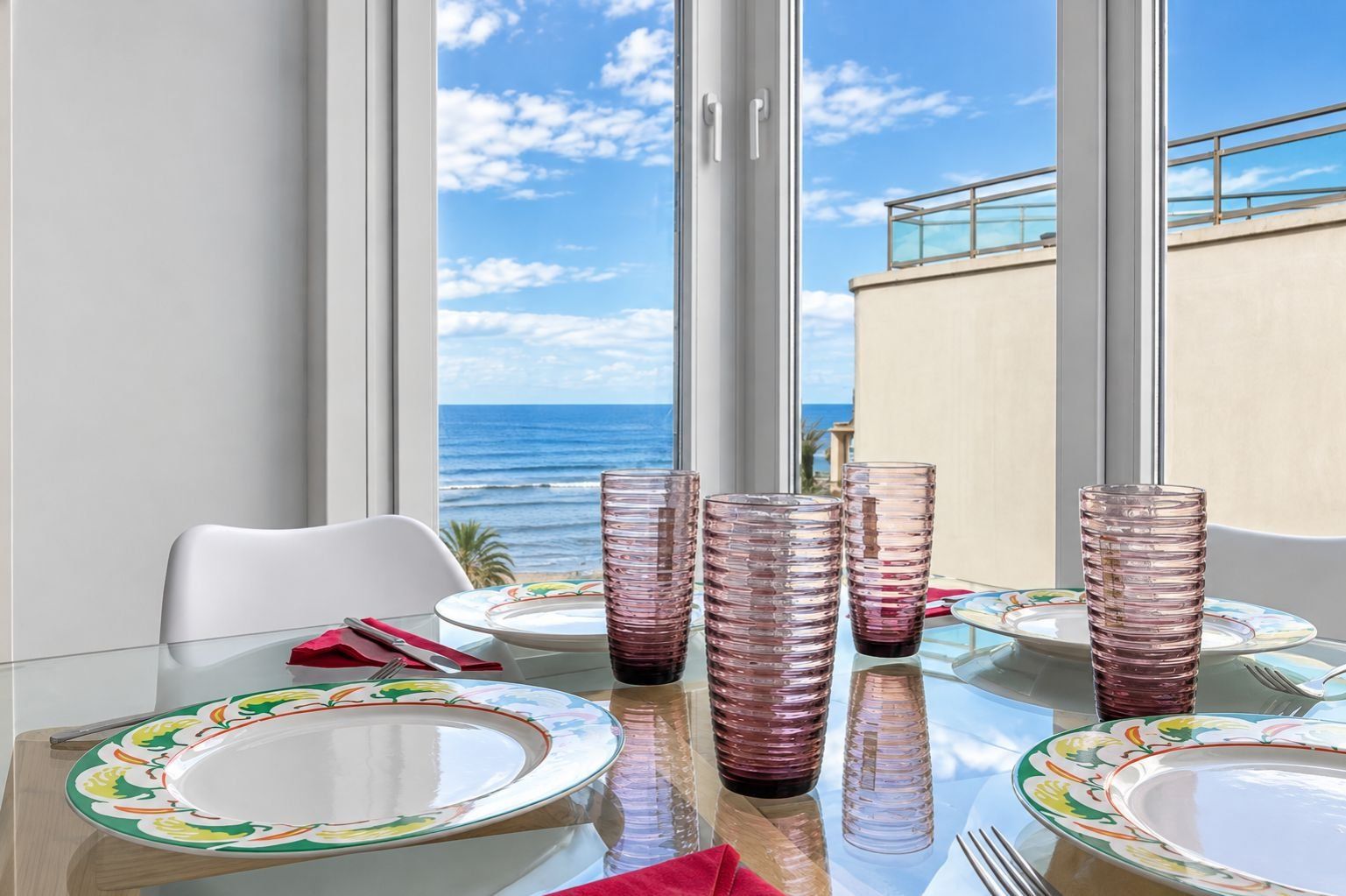 A dining table with plates, cutlery, and pink glassware set against a backdrop of a clear sky and ocean view.