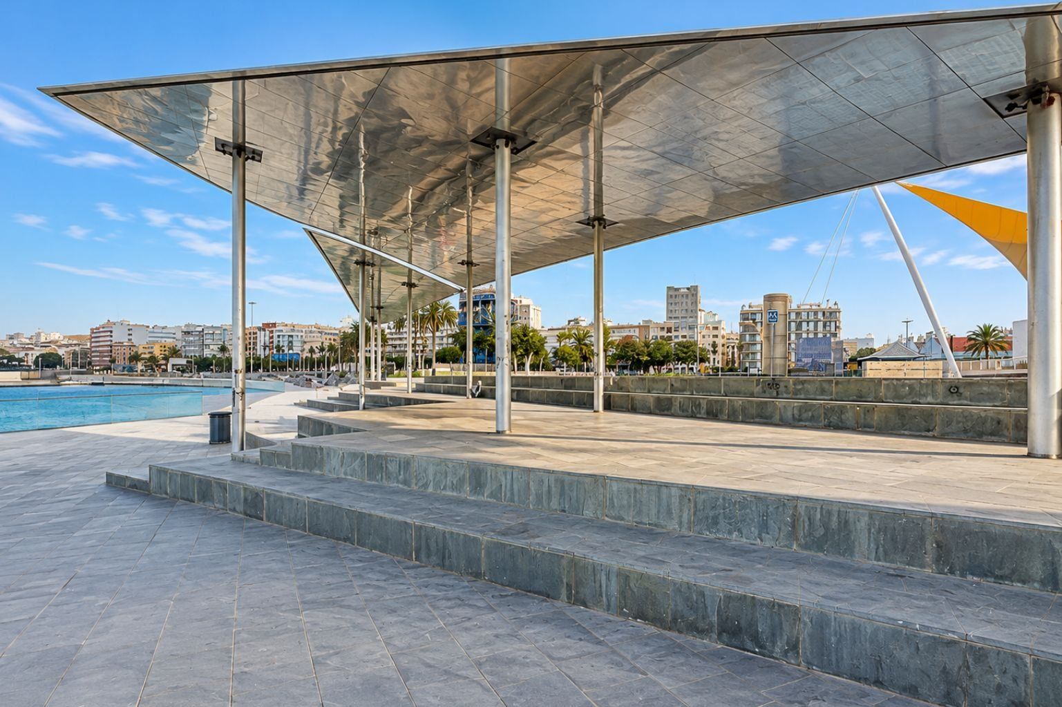 A waterfront promenade with a covered seating area, steps leading to a pool, and a cityscape in the background.